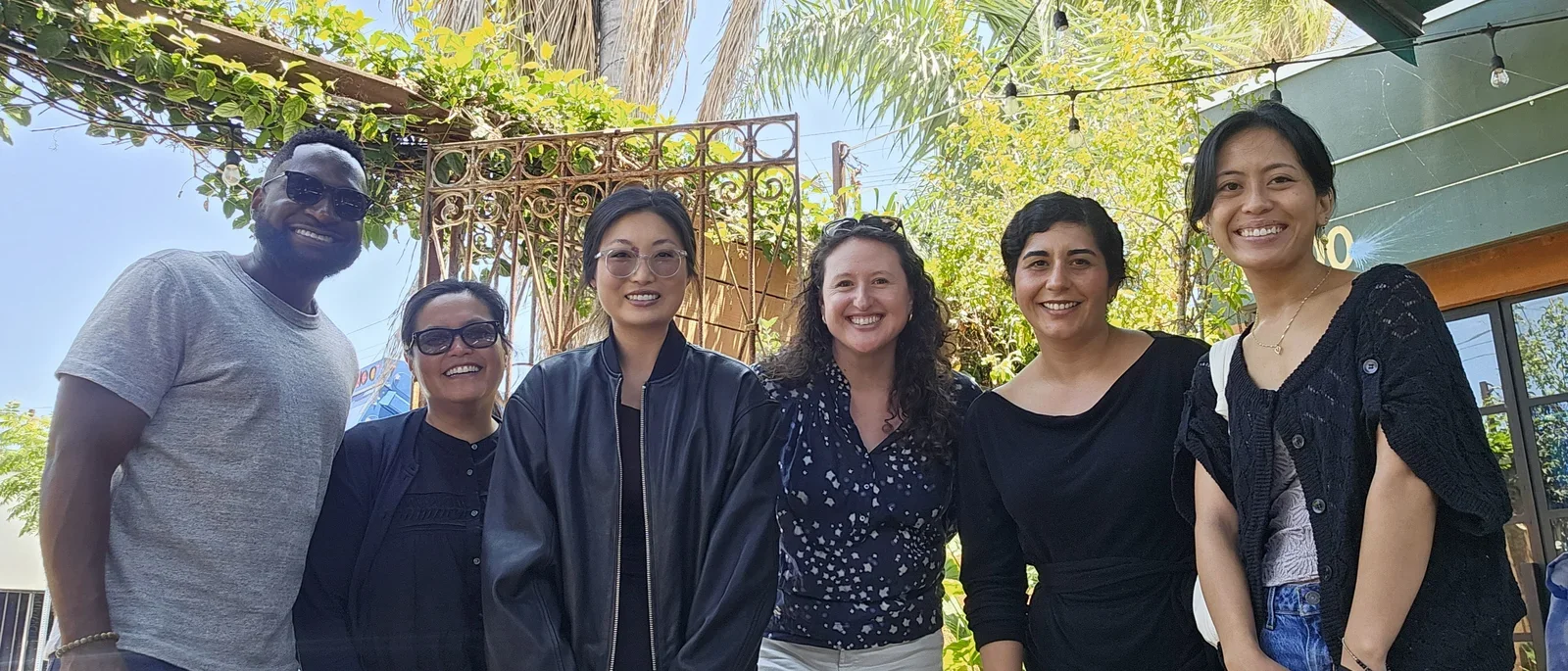 6 people standing in a row smiling with trees and gate in the background