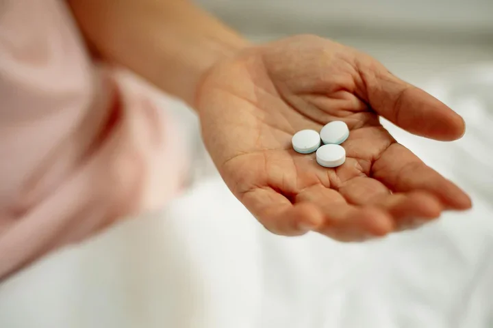 Hand with three white pill of person with a pink shirt in the background