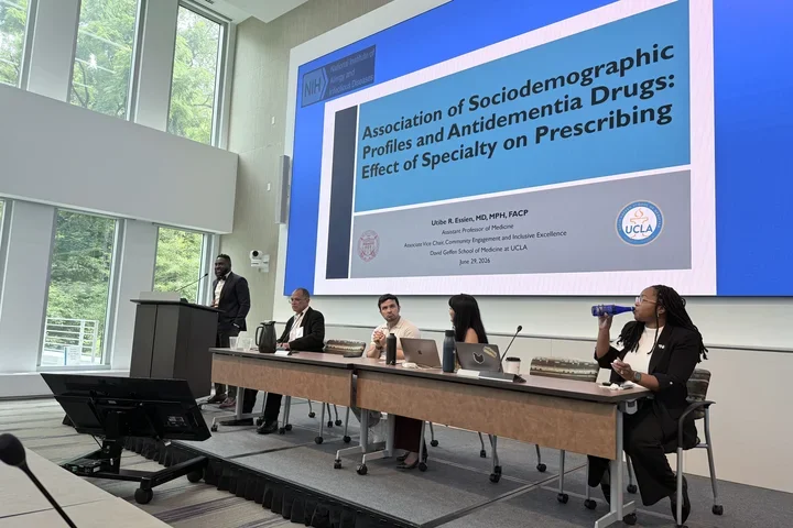 One man at the podium with 4 people seated at a panel table with slide of presentation in the background