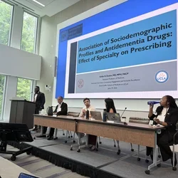One man at the podium with 4 people seated at a panel table with slide of presentation in the background